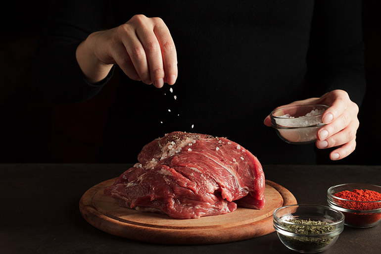 Imagem de uma carne vermelha sob uma tábua de carne com uma mão acima salpicando sal grosso sob ela.