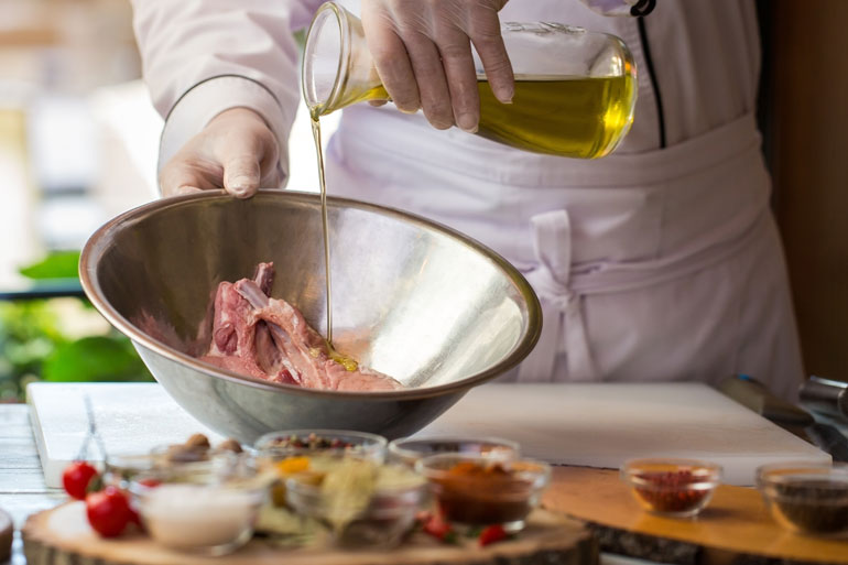 A imagem mostra um chefe de cozinha colocando azeite em uma carne bovina que está em uma tigela de alumínio.