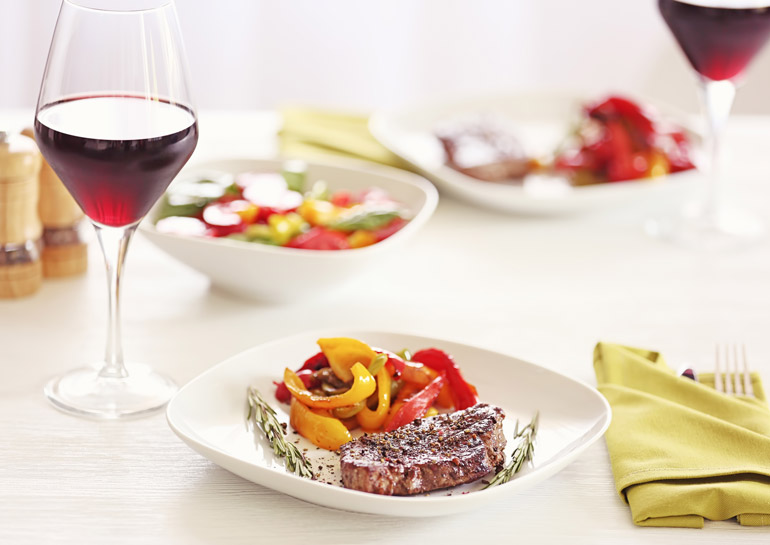 Taça com vinho até a metade sob uma mesa ao lado de um prato branco com um pedaço de carne e legumes. 