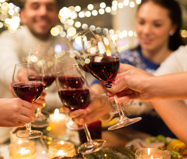 Braços de pessoas com taças de vinho nas mãos fazendo um brinde sentados a uma mesa decorada com tema de final de ano.