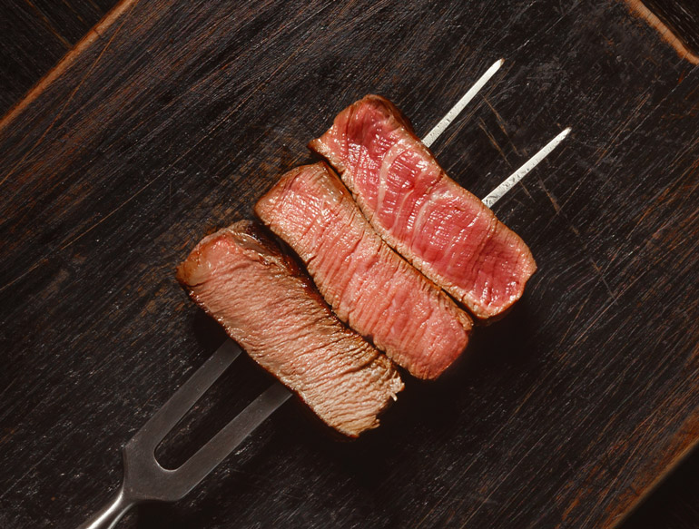 Espeto de carnes sob uma mesa de madeira com três pedaços pequenos de carnes em pontos diferentes, um malpassado, outro ao ponto e um terceiro bem passado.